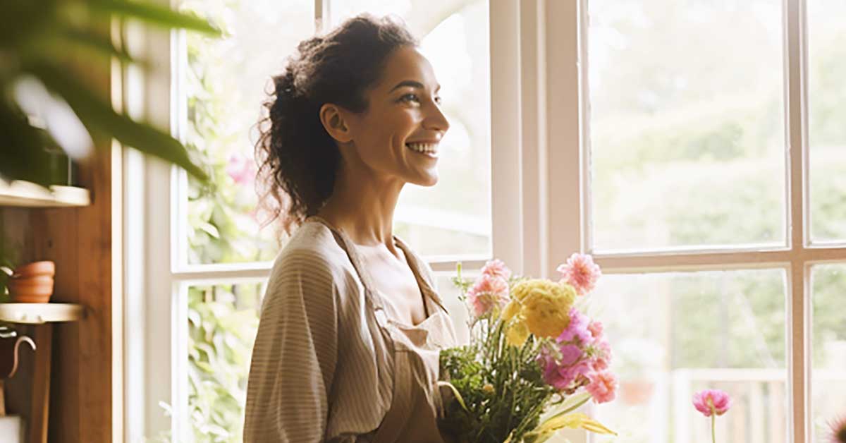 Woman smiling and holding a bunch of flowers while standing next to a window on a sunny summer day.