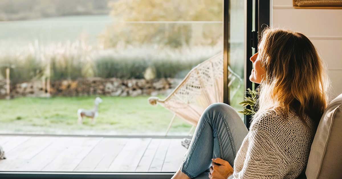 Woman looking out open door at sunny country landscape while she relaxes after downsizing her home.