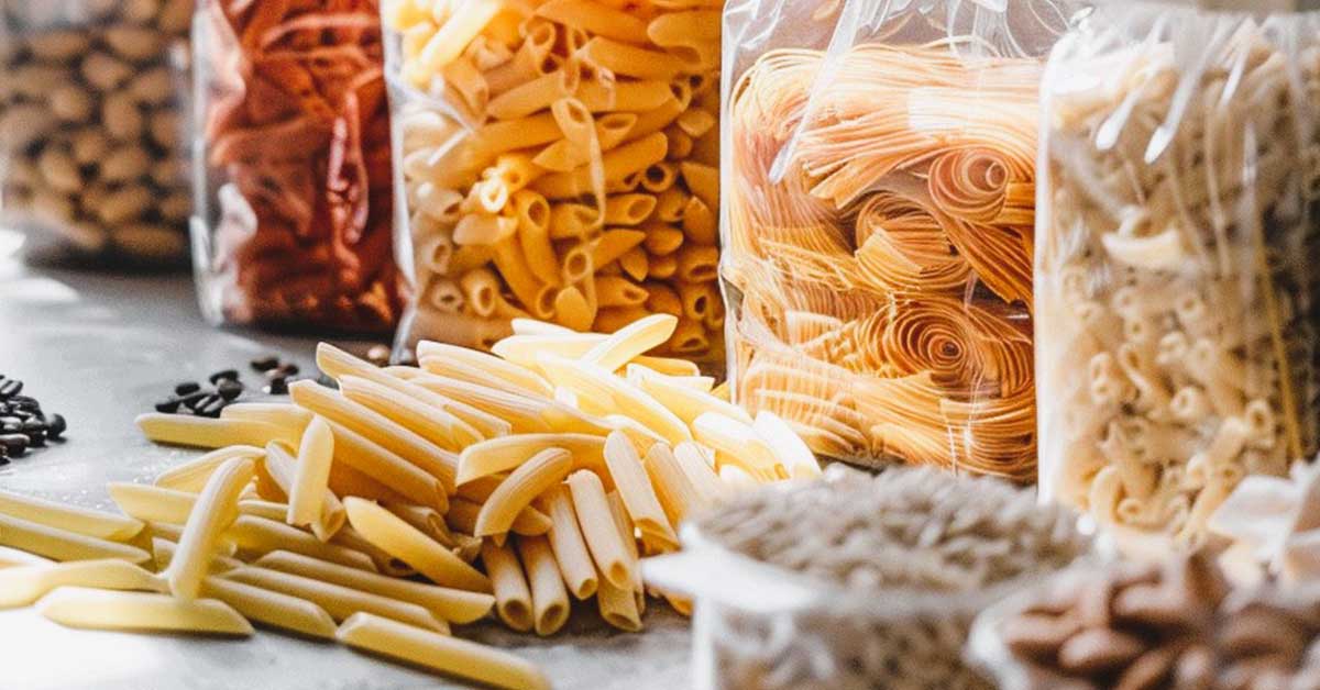 Various types of pasta from a large stockpile, including penne, rotini, and spaghetti, stored in clear plastic bags and displayed on a countertop.