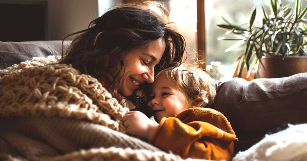A mother and her child share a simple and cozy moment, wrapped in blankets and smiling warmly at each other, while sitting on a couch near a sunlit window with a green plant in the background.