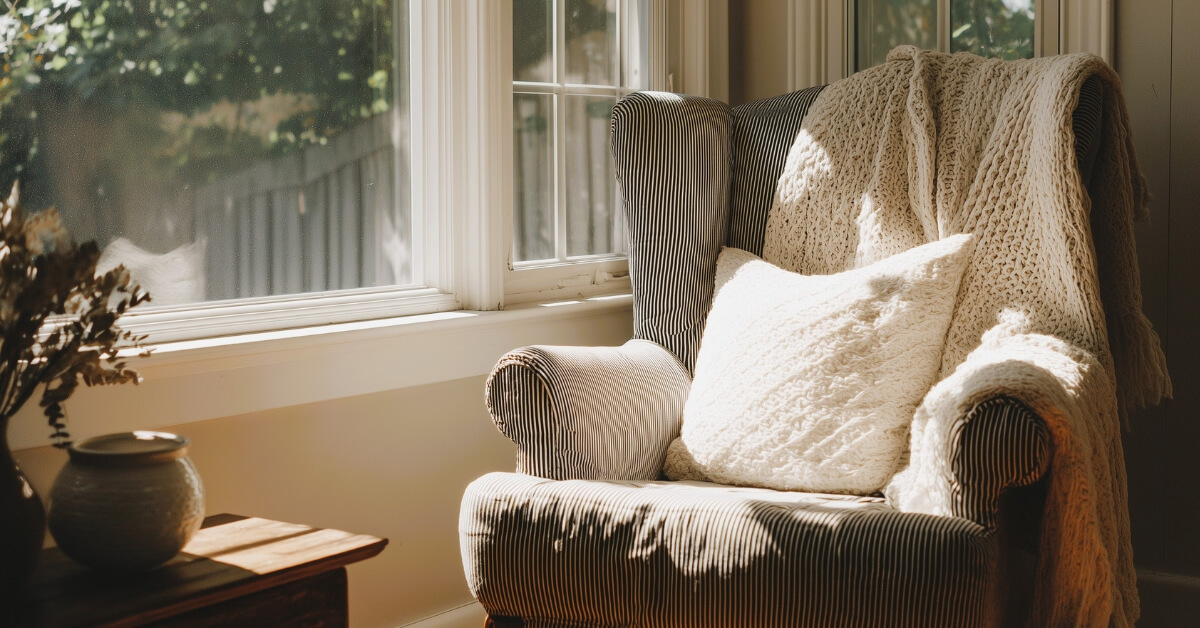 Sunlit cozy armchair with a knit blanket and pillow beside a window, creating a warm minimalist home setting that reflects low spend month rules by emphasizing simple comforts and enjoying what you already have.