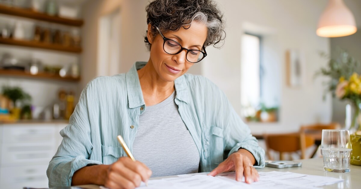 Woman with short curly hair and glasses sits at a kitchen table while planning meals and organizing a reusable grocery list. Bright kitchen shelves and a glass of water sit in the background creating a calm home planning scene.