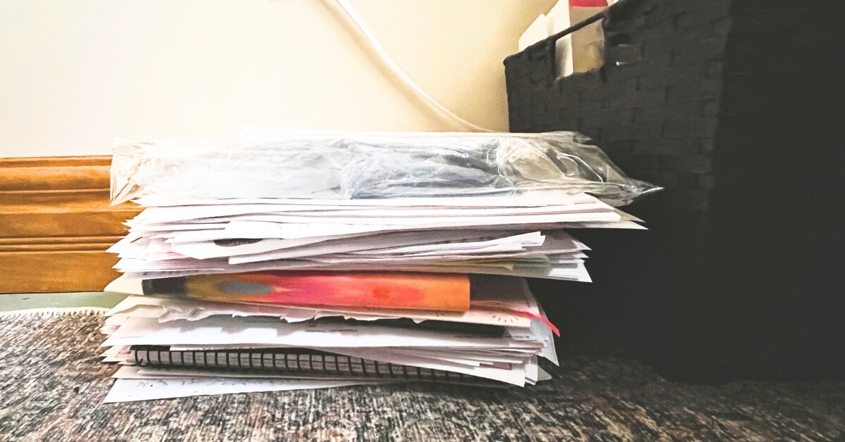 Stack of mixed papers, envelopes, and notebooks piled on the floor beside a black basket, showing the need for learning how to declutter paper piles.